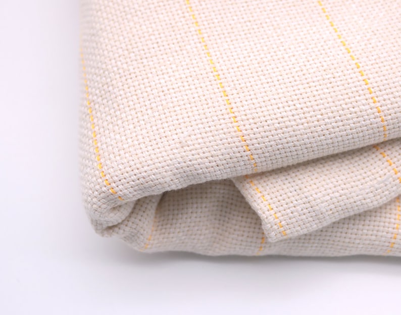 May include: A close-up of a folded piece of off-white fabric with thin yellow stripes. The fabric is woven and has a slightly textured surface.