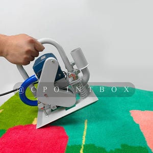 May include: A hand holds a gray and blue carpet cleaning machine with a blue hose and a spring. The machine is on a green and pink carpet with a floral pattern.