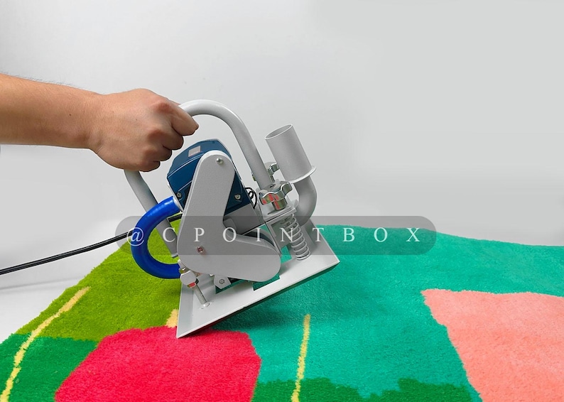 May include: A hand holds a gray and blue carpet cleaning machine with a blue hose and a spring. The machine is on a green and pink carpet with a floral pattern.