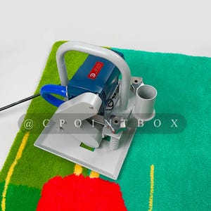 May include: A blue and gray electric carpet cleaning machine with a handle and a hose. The machine is on a green and red carpet with a yellow stripe.
