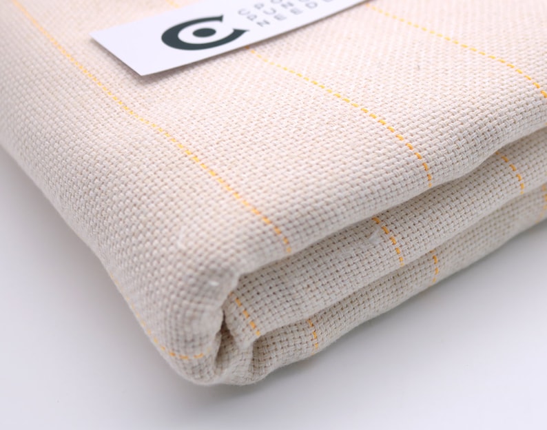 May include: A close-up of a folded piece of off-white fabric with thin yellow stripes. The fabric is woven and has a textured surface.