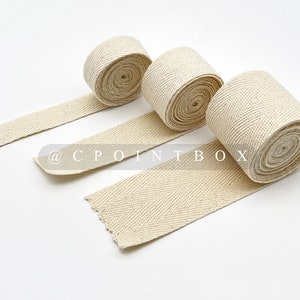 May include: Three rolls of natural-colored, woven cotton ribbon with a herringbone pattern. The ribbon is unspooled, revealing the texture and weave. The text "@ CPOINTBOX" is visible on the ribbon.
