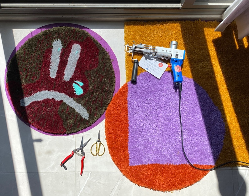 K&ouml;nnte beinhalten: Zwei runde, farbenfrohe, getuftete Teppiche mit einem lila und orangefarbenen Teppich im Vordergrund und einem roten, gr&uuml;nen und grauen Teppich im Hintergrund. Ein blaues und wei&szlig;es Werkzeug mit einem schwarzen Kabel liegt auf dem lila Teppich. Zwei Paar rote und silberne Scheren liegen auf der wei&szlig;en Oberfl&auml;che.