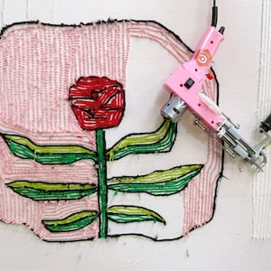 May include: A pink and white electric rug hooking machine with a black handle. The machine is hooked to a white canvas with a red flower and green leaves embroidered on it.