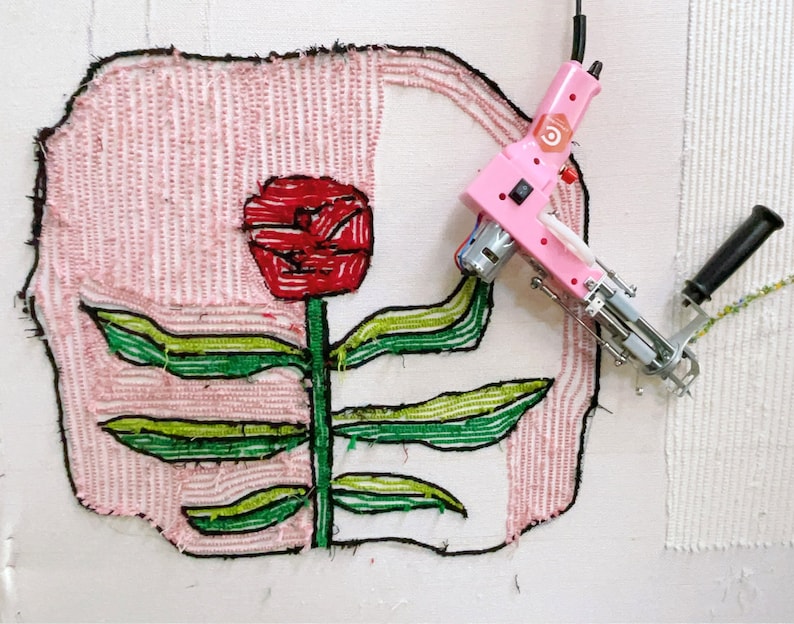 May include: A pink and white electric rug hooking machine with a black handle. The machine is hooked to a white canvas with a red flower and green leaves embroidered on it.