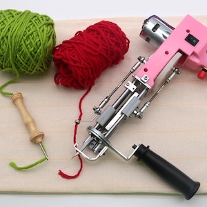 May include: A pink electric rug hooking machine with a black handle, a green ball of yarn, a red ball of yarn, and a wooden yarn needle on a beige striped surface.