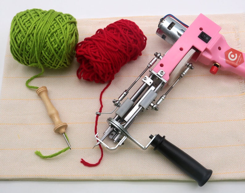 May include: A pink electric rug hooking machine with a black handle, a green ball of yarn, a red ball of yarn, and a wooden yarn needle on a beige striped surface.