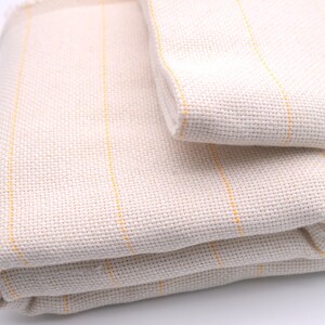 May include: A white fabric with thin yellow stripes folded in thirds. The fabric is woven and has a textured surface.