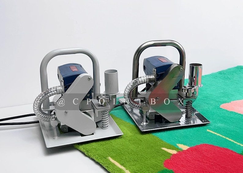 May include: Two gray and blue electric carpet tufting machines with handles and a clear hose. The machines are on a green and red patterned carpet.