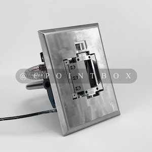 May include: A silver metal wall plate with a rectangular cutout and a small, silver, rectangular device inside. The device has a series of small, black, rectangular buttons. The plate is mounted on a white wall.
