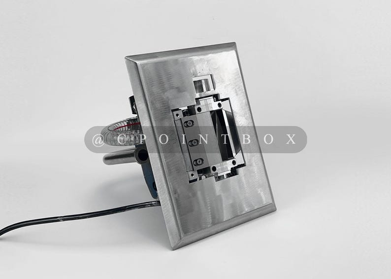 May include: A silver metal wall plate with a rectangular cutout and a small, silver, rectangular device inside. The device has a series of small, black, rectangular buttons. The plate is mounted on a white wall.