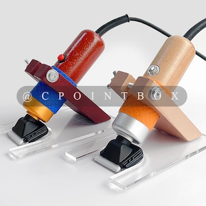 Puede incluir: Dos cortadoras eléctricas con mangos de madera y bases de acrílico transparente. Las cortadoras están sobre una superficie blanca. Las cortadoras tienen cables de diferentes colores y agarres de goma de diferentes colores. Las cortadoras están etiquetadas con el texto "@CPOINTBOX".