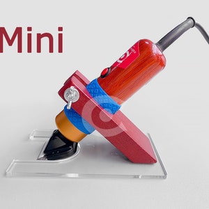 Könnte beinhalten: Ein rotes, hölzernes Mini-Elektro-Schnitzwerkzeug mit blau-weißem Klebeband am Griff, das auf einem durchsichtigen Acrylsockel ruht. Das Werkzeug ist eingesteckt und einsatzbereit. Das Wort "Mini" steht in roten Buchstaben über dem Werkzeug.