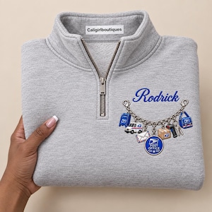 Könnte beinhalten: Ein hellgraues Sweatshirt mit kurzem Reißverschluss und silbernem Zipper sowie einem Grafikdruck auf der Brust. Das Design zeigt den Namen Rodrick in blauer Schrift über einem silbernen Bettelarmband mit Post-Motiven.