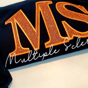 Pu&ograve; includere: Felpa blu navy con le lettere ricamate "MS" in arancione glitterato e le parole "Multiple Sclerosis" in corsivo bianco. &Egrave; presente anche un nastro arancione ricamato.