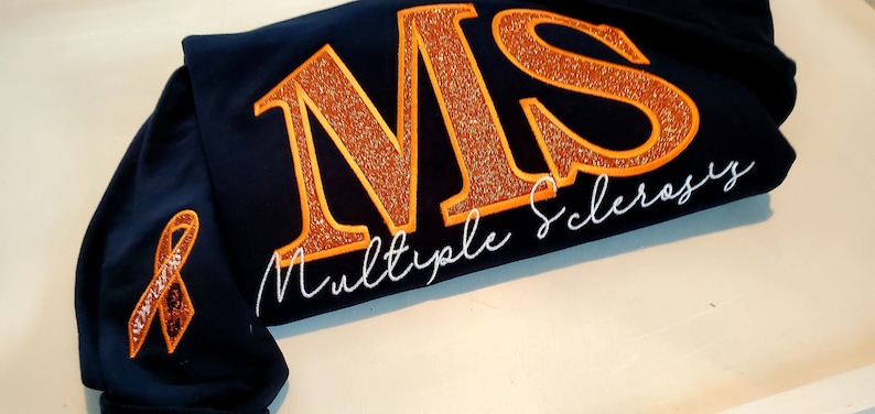 Pu&ograve; includere: Felpa blu navy con le lettere ricamate "MS" in arancione glitterato e le parole "Multiple Sclerosis" in corsivo bianco. &Egrave; presente anche un nastro arancione ricamato.