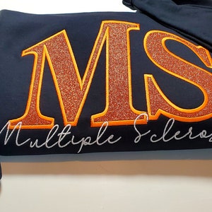Pu&ograve; includere: Felpa blu navy con le lettere "MS" in glitter arancione, bordate di arancione. Le parole "Multiple Sclerosis" sono ricamate in corsivo bianco. Un nastro arancione con testo &egrave; sulla manica.