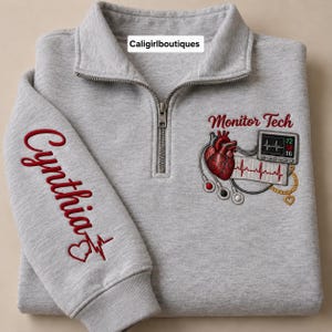 May include: A light gray quarter-zip sweatshirt featuring embroidered designs. The sleeve displays the name Cynthia in red with a heart EKG line. The chest shows a red anatomical heart, an EKG monitor, and the text Monitor Tech in red.