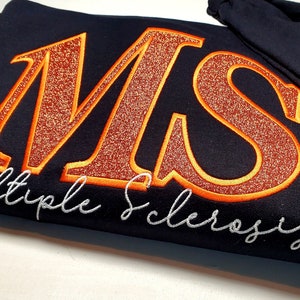 Pu&ograve; includere: Una felpa blu navy con le lettere ricamate "MS" in glitter arancione con un contorno arancione. Le parole "Multiple Sclerosis" sono ricamate in corsivo bianco sotto. Un nastro arancione &egrave; ricamato sulla manica.