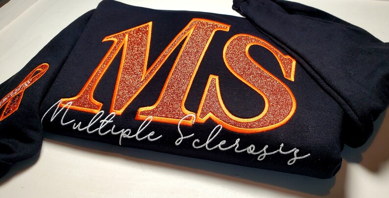 Pu&ograve; includere: Una felpa blu navy con le lettere ricamate "MS" in glitter arancione con un contorno arancione. Le parole "Multiple Sclerosis" sono ricamate in corsivo bianco sotto. Un nastro arancione &egrave; ricamato sulla manica.