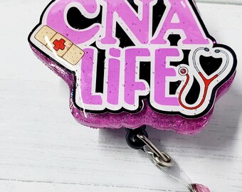 May include: A pink glitter badge reel with the words "CNA LIFE" in a bubble letter style. The badge features a bandage and a stethoscope. The reel has a black clip and a clear plastic strap with a pink gem.