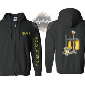 May include: Black zip-up hoodie with "NAME" in yellow on the chest and "HOUSEKEEPING" down the sleeve. The back features a cleaning cart graphic with a mop, bucket, and cleaning supplies. A practical and comfortable garment.