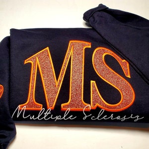Pu&ograve; includere: Felpa blu navy con le lettere "MS" in glitter arancione, bordate di arancione, e le parole "Multiple Sclerosis" in corsivo bianco. Un nastro arancione &egrave; sulla manica.