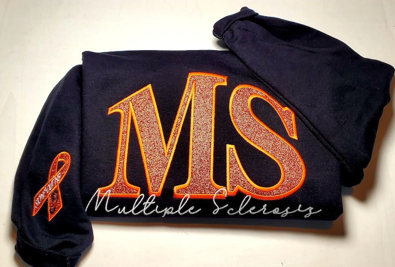 Pu&ograve; includere: Felpa blu navy con le lettere "MS" in glitter arancione, bordate di arancione, e le parole "Multiple Sclerosis" in corsivo bianco. Un nastro arancione &egrave; sulla manica.