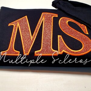 Pu&ograve; includere: Felpa con cappuccio nera con le lettere "MS" in glitter arancione, bordate d'oro, e le parole "Multiple Sclerosis" in corsivo bianco. Un nastro arancione di sensibilizzazione &egrave; sulla manica.