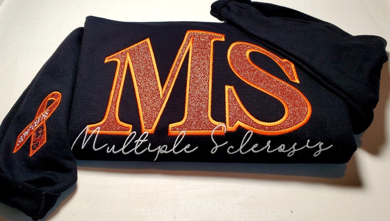 Pu&ograve; includere: Felpa con cappuccio nera con le lettere "MS" in glitter arancione, bordate d'oro, e le parole "Multiple Sclerosis" in corsivo bianco. Un nastro arancione di sensibilizzazione &egrave; sulla manica.