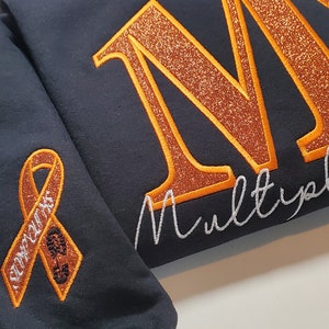 Pu&ograve; includere: Felpa blu navy scuro con lettere glitterate arancioni che compongono "MS" e le parole "Multiple Sclerosis" in corsivo bianco. Un nastro arancione con la scritta "MS Awareness" &egrave; sulla manica.
