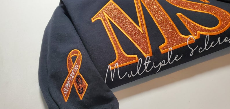 Pu&ograve; includere: Felpa blu navy scuro con lettere glitterate arancioni che compongono "MS" e le parole "Multiple Sclerosis" in corsivo bianco. Un nastro arancione con la scritta "MS Awareness" &egrave; sulla manica.