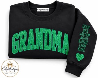 Op de afbeelding: Een zwart sweatshirt met ronde hals en het woord GRANDMA in grote, gebogen, glinsterende groene letters op de borst. De mouw toont een lijst met namen waaronder Leah, Bea, Jayden, Noah, Kenzie, Leon en Kobe boven een groen glitterhart.