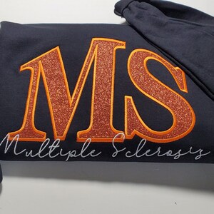 Pu&ograve; includere: Una felpa blu navy con le lettere "MS" in grandi lettere arancioni glitterate. Le parole "Multiple Sclerosis" sono scritte sotto in corsivo. Un nastro arancione &egrave; ricamato sulla manica.