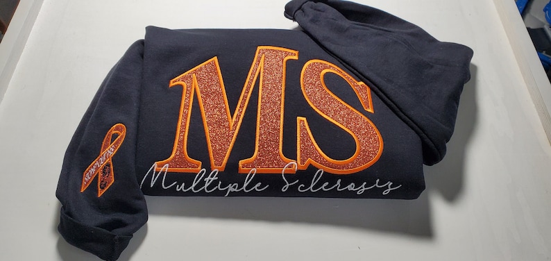 Pu&ograve; includere: Una felpa blu navy con le lettere "MS" in grandi lettere arancioni glitterate. Le parole "Multiple Sclerosis" sono scritte sotto in corsivo. Un nastro arancione &egrave; ricamato sulla manica.