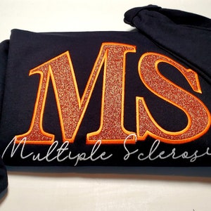 Pu&ograve; includere: Una felpa con cappuccio blu navy con le lettere ricamate "MS" in glitter arancione, bordate di arancione. Le parole "Multiple Sclerosis" sono ricamate in corsivo bianco sotto. Un nastro arancione di sensibilizzazione &egrave; sulla manica.