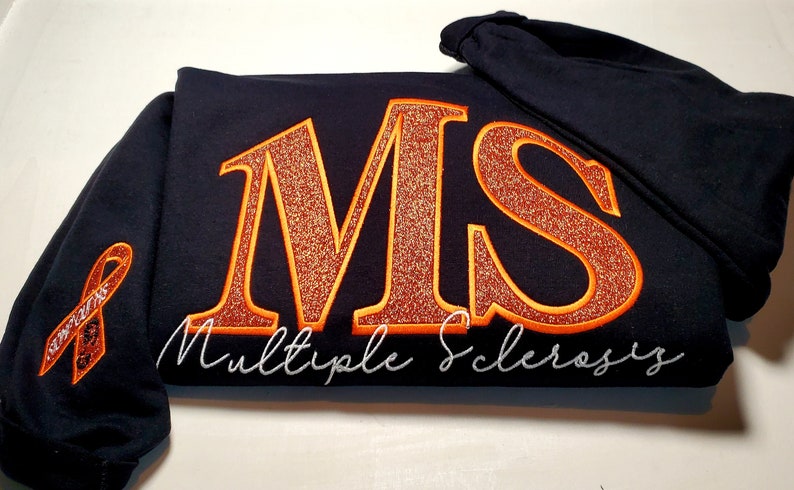 Pu&ograve; includere: Una felpa con cappuccio blu navy con le lettere ricamate "MS" in glitter arancione, bordate di arancione. Le parole "Multiple Sclerosis" sono ricamate in corsivo bianco sotto. Un nastro arancione di sensibilizzazione &egrave; sulla manica.