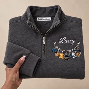 Könnte beinhalten: Ein anthrazitfarbenes Sweatshirt mit kurzem Reißverschluss und einem weißen Etikett mit der Aufschrift Caligirlboutiques. Auf der Brust steht der Name Larry in weißer Schreibschrift über einer Grafik eines Amazon-Charm-Armbands.