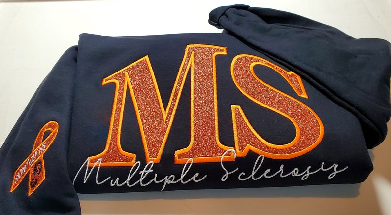 Pu&ograve; includere: Felpa con cappuccio blu navy con le lettere "MS" in glitter arancione, bordate di arancione, e le parole "Multiple Sclerosis" in corsivo bianco. Un nastro arancione di sensibilizzazione &egrave; sulla manica.