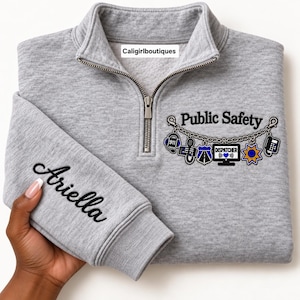May include: A heather grey quarter-zip sweatshirt featuring a silver zip and a graphic on the chest that reads Public Safety with a chain design holding icons of a headset, badge, computer, and phone. The sleeve is embroidered with the name Ariella.