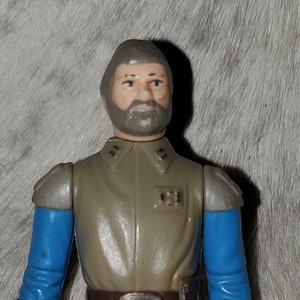 Op de afbeelding: Een vintage Star Wars-actionfiguur van een rebellenofficier met een grijs uniform, blauwe mouwen en een zwarte riem. De figuur heeft een grijze baard en een serieuze uitdrukking.