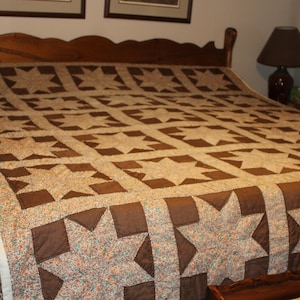 May include: A large bed quilt featuring a repeating pattern of eight-pointed stars. The stars are constructed from a light-colored fabric with a small floral print, while the surrounding blocks and sashing are made of solid dark brown fabric.