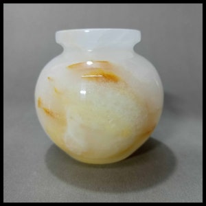 May include: A small, white alabaster vase with a round body and a slightly flared opening. The vase has a swirling pattern of yellow and orange throughout.