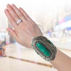 May include: A silver cuff bracelet featuring a large, rectangular, bright green stone. The bracelet has an ornate, engraved design. A silver ring with three clear stones is also visible.
