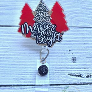 Op de afbeelding: Een rode en zwarte glitter kerstboomvormige badgehouder met de tekst "Merry & Bright" in goudglitter. De badgehouder heeft een zwarte glitterbedel bevestigd aan de intrekbare badgehaspel.