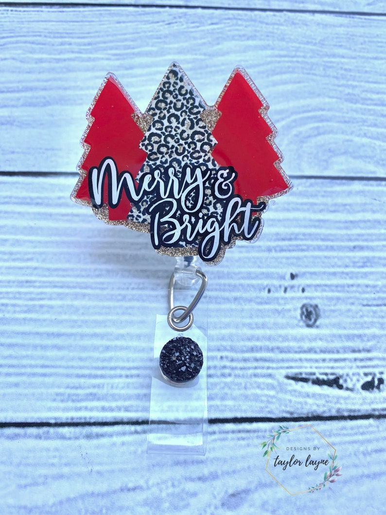 Op de afbeelding: Een rode en zwarte glitter kerstboomvormige badgehouder met de tekst "Merry & Bright" in goudglitter. De badgehouder heeft een zwarte glitterbedel bevestigd aan de intrekbare badgehaspel.