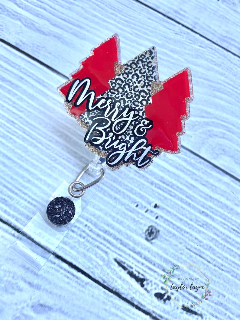 Op de afbeelding: Een rode en zwarte kerstboomvormige badgehouder met de tekst "Merry & Bright" in gouden glitter. De badgehouder heeft een zwarte glitterbedel bevestigd aan een transparante intrekbare badgehaspel.