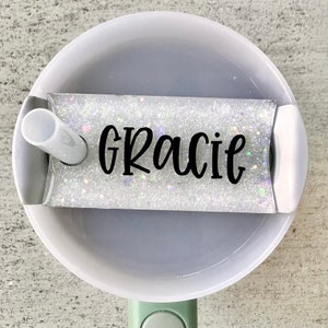 May include: A white glitter tumbler with a white lid and a straw. The tumbler has the name "Gracie" written in black lettering.