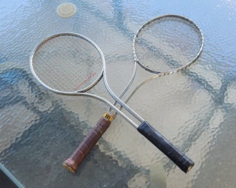 May include: Two silver badminton rackets with black and brown grips. The rackets are on a glass table.
