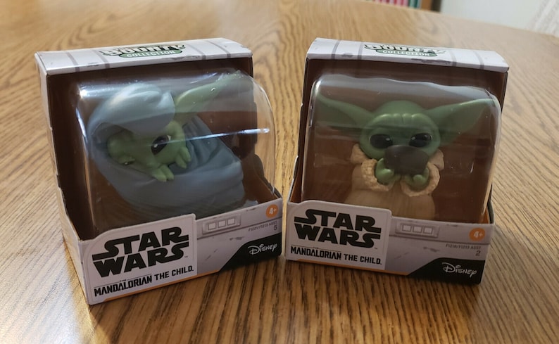 May include: Two unopened Star Wars Mandalorian The Child Bounty Collection figures. The figures are in plastic packaging and are both green. One figure is sitting in a gray pouch and the other is holding a small black object.